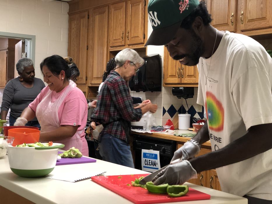 Man cutting green bell pepper on red board; people preparing food in kitchen; "HELLO CLEAN" sign