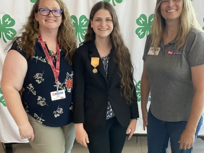 Three people standing in front of 4‑H clover backdrop; center person wears a medal