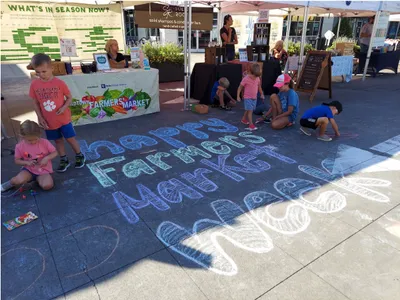 Children drawing chalk "Happy Farmers Market" on pavement in front of outdoor market stalls