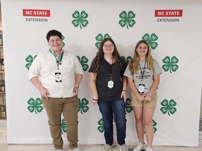 Three teens wearing 4-H badges standing before NC STATE EXTENSION backdrop with green 4‑H clovers
