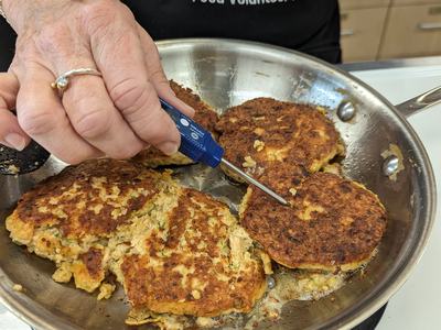 salmon burger patties being cooked in a skillet on a stove. Extension Master Food Volunteer is checking the temperature of the patties with a thermometer.