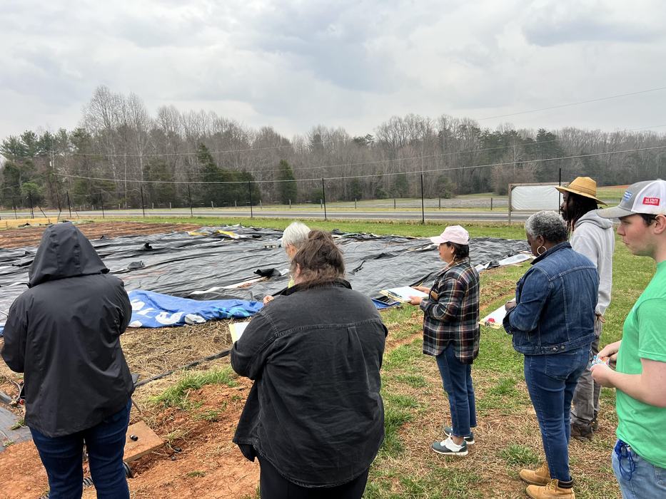 Group of people standing at edge of tarped field, some holding clipboards