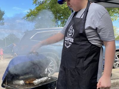 Teen in blue cap and "Grill Master" apron lifting grill lid with smoking food