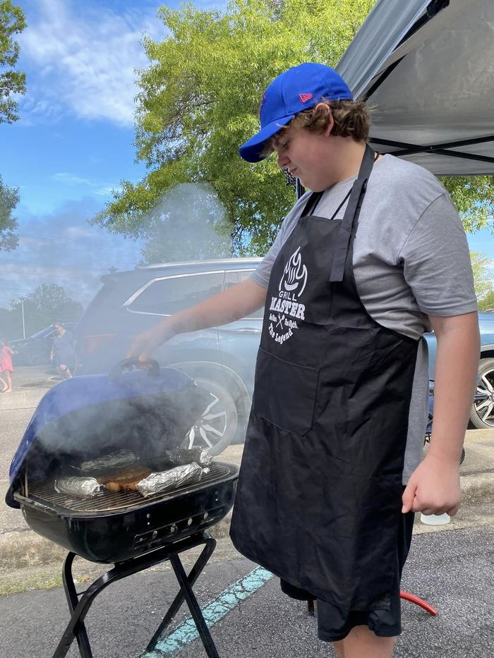 A boy in a Grill Master apron inspects a grill with food.