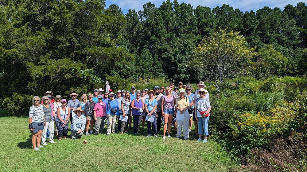 Large group of people standing in a grassy clearing by trees, many wearing hats