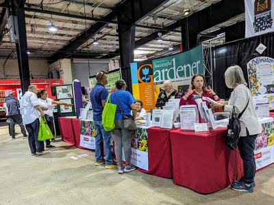 Booth for "Successful Gardener" with staff advising visitors at an indoor fair
