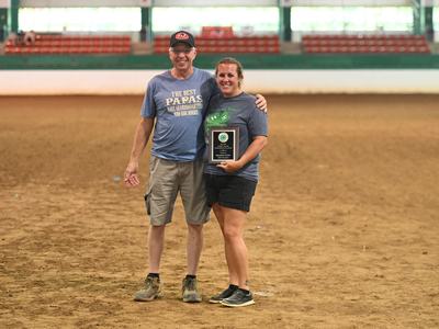 Man and woman standing in an arena, woman holding a plaque award.