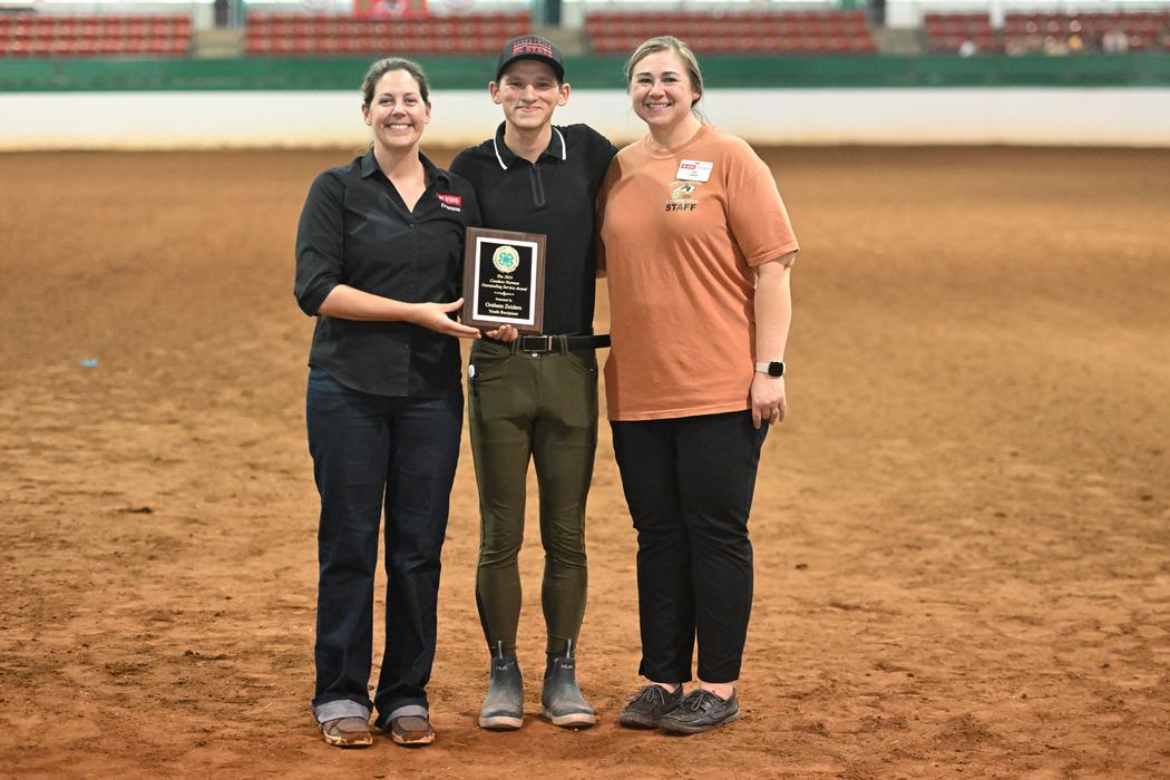 Three people standing in an arena holding a plaque with a 4‑H emblem