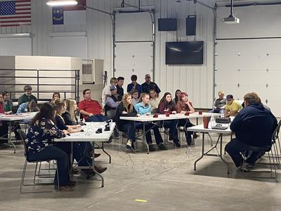 People seated around folding tables in a garage-style room for a meeting.