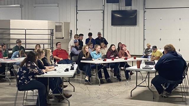 People seated around folding tables in a garage-style room for a meeting.