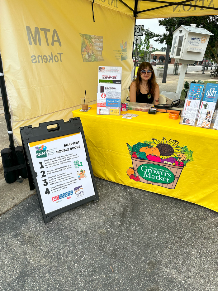 Woman seated at yellow Growers Market booth with SNAP/EBT "Double Bucks" sign