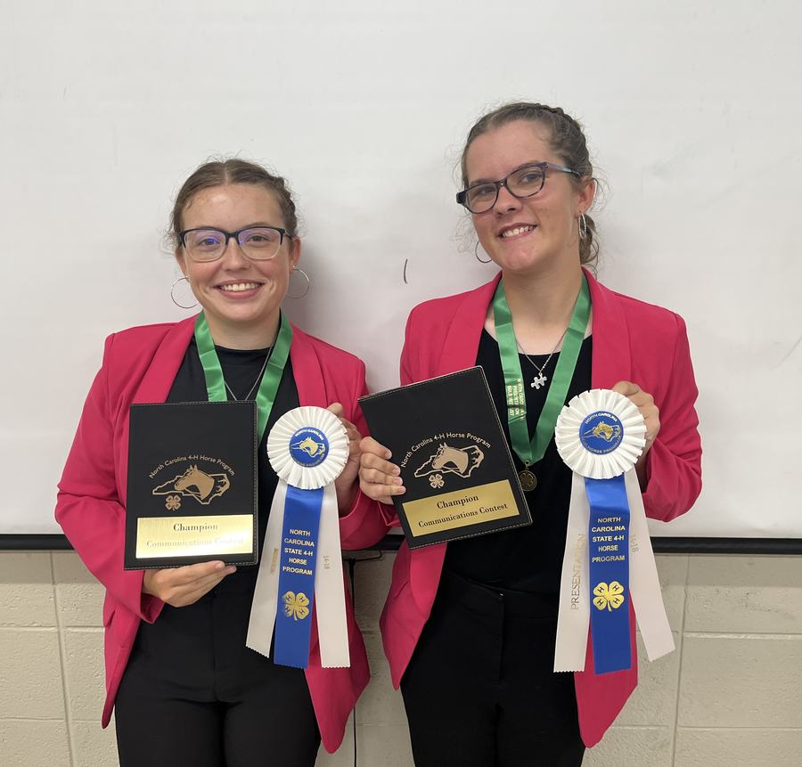 Two people in pink jackets holding "Champion" plaques and North Carolina State 4‑H Horse Program ribbons