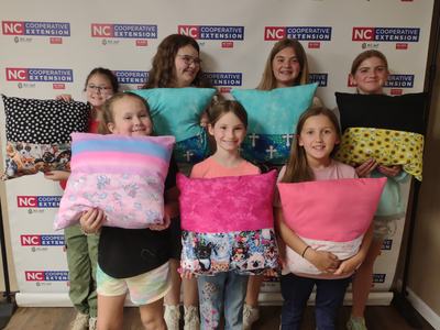 Seven girls holding handmade pillows in front of NC Cooperative Extension backdrop