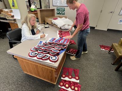 Two people at a desk sorting red and blue award ribbons; wall sign reads "AGRICULTURAL DISTRICT"