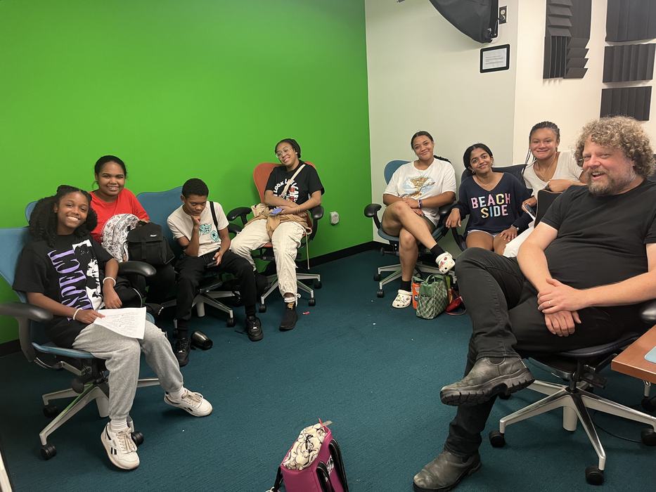 Group of eight people seated in office chairs against a green screen wall