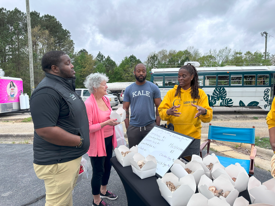 Four people at outdoor market stall; vendor in yellow hoodie explaining over boxes of mushrooms.