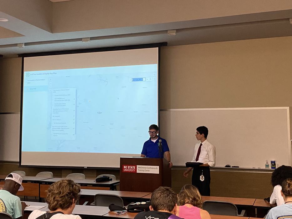 Two presenters at podium in lecture hall with projected screen; podium sign "NC STATE UNIVERSITY"