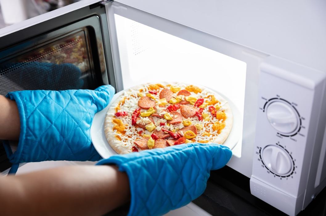 pizza going into microwave with blue potholders on hands
