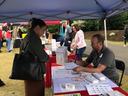 Person at outdoor vendor tent table speaking with seated man handling paperwork