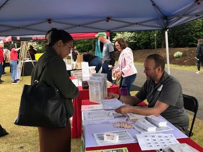 Person at outdoor vendor tent table speaking with seated man handling paperwork