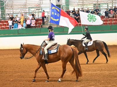 Two riders on horses carry a North Carolina flag reading "NC" and a white 4‑H clover flag.