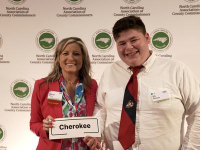 Two people at North Carolina Association of County Commissioners backdrop holding sign "Cherokee"