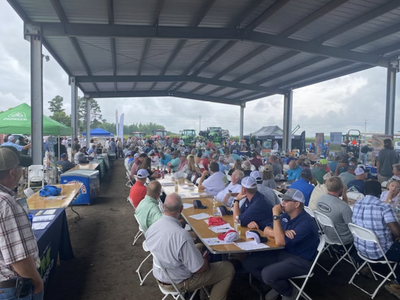 Large crowd seated at long tables under a pavilion at an outdoor agricultural equipment expo