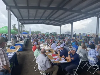 Large crowd seated at long tables under a pavilion at an outdoor agricultural equipment expo