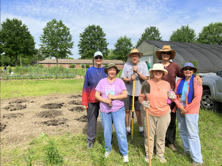 Six people standing in a community garden holding shovels and rakes in front of planting plots.