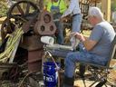 Three men feed sugarcane into a vintage cane press under a shed; blue bucket labeled Lowe's.