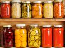 Subject: Horizontal view of three wooden shelves holding a variety of canned vegetables and fruits, lined up in rows of glass jars. Food staples canned include sauces, tomatoes, corn, sweet potatoes, sauerkraut, dill pickles, red beets, peaches, grape jel
