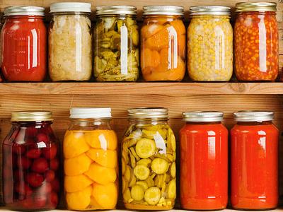 Subject: Horizontal view of three wooden shelves holding a variety of canned vegetables and fruits, lined up in rows of glass jars. Food staples canned include sauces, tomatoes, corn, sweet potatoes, sauerkraut, dill pickles, red beets, peaches, grape jel