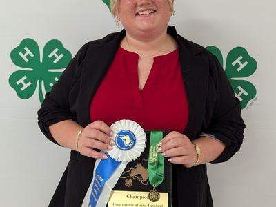 Person holding plaque reading "Champion Communications Contest" with ribbons and 4‑H clover backdrop