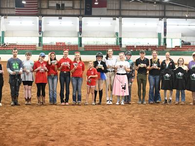 Group of young riders and participants lined up in indoor arena holding awards