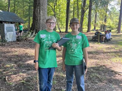 Two boys in green shirts holding clipboards standing under a large tree in a wooded park