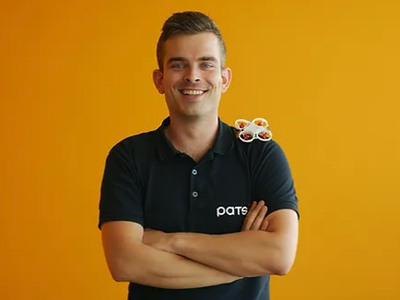 Man with arms crossed in black polo labeled "pats", small toy cat on his shoulder