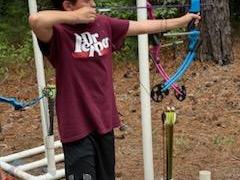 Boy wearing "Dr Pepper" shirt draws a blue recurve bow at an outdoor archery setup