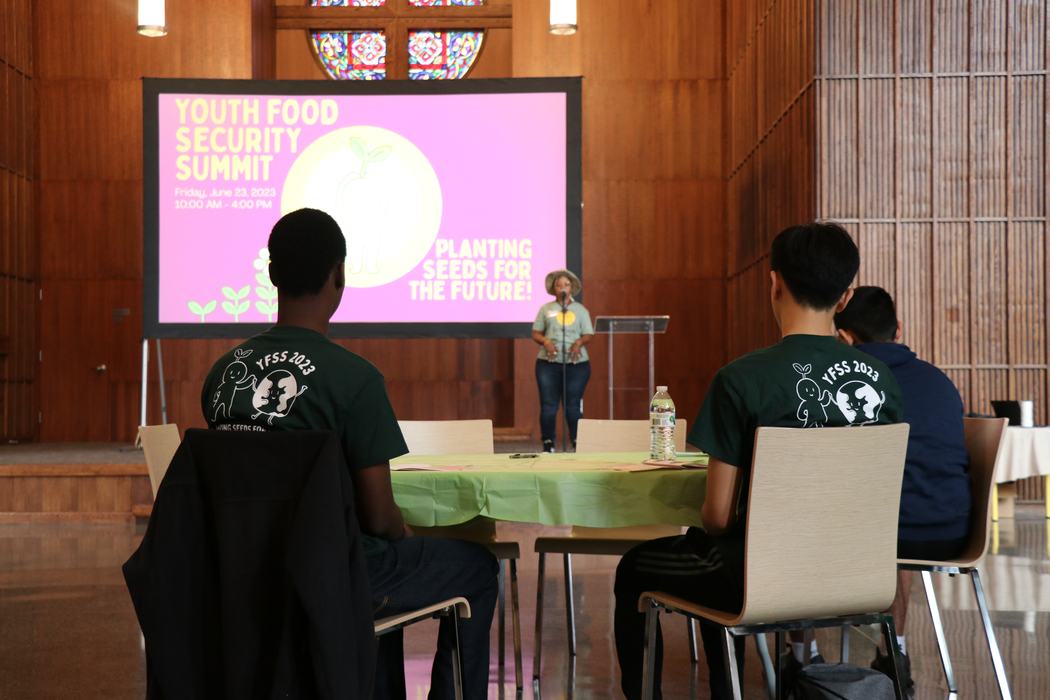 A woman gives a talk to kids sitting at tables.