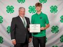 Man in green polo holds certificate reading "National 4‑H Conference Delegate Noah Snyder" beside man in suit
