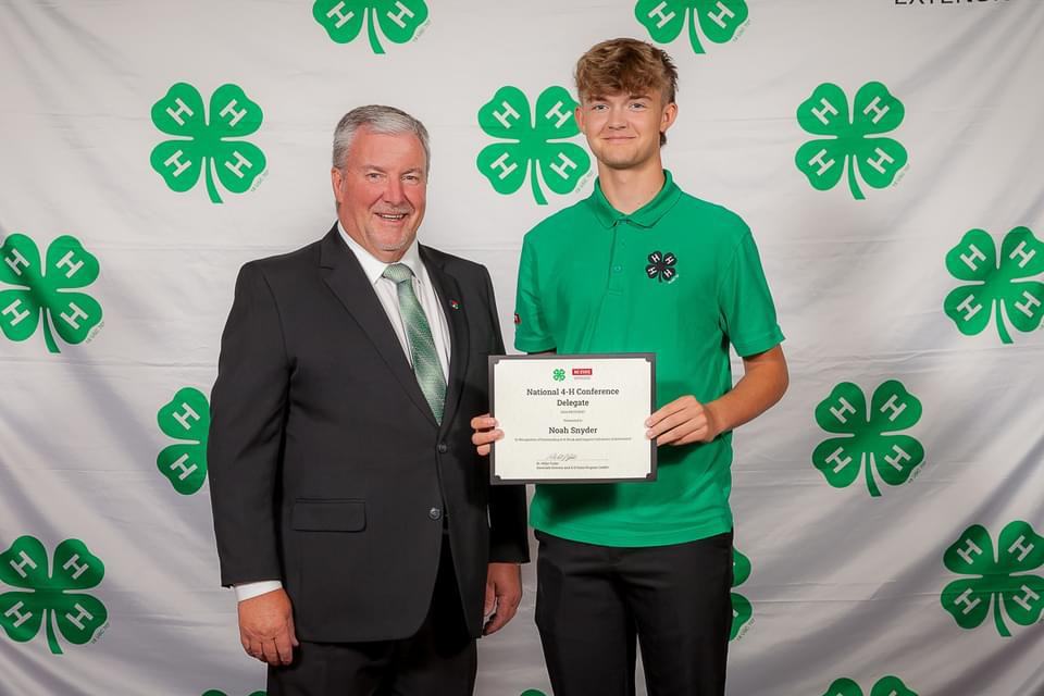 Man in green polo holds certificate reading "National 4‑H Conference Delegate Noah Snyder" beside man in suit