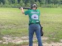 Person wearing green jersey labeled "ROBERTSON" and "LEARN BY DOING" aiming a shotgun at an outdoor range