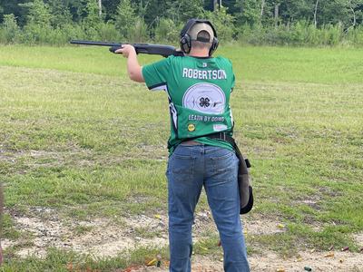 Person wearing green jersey labeled "ROBERTSON" and "LEARN BY DOING" aiming a shotgun at an outdoor range