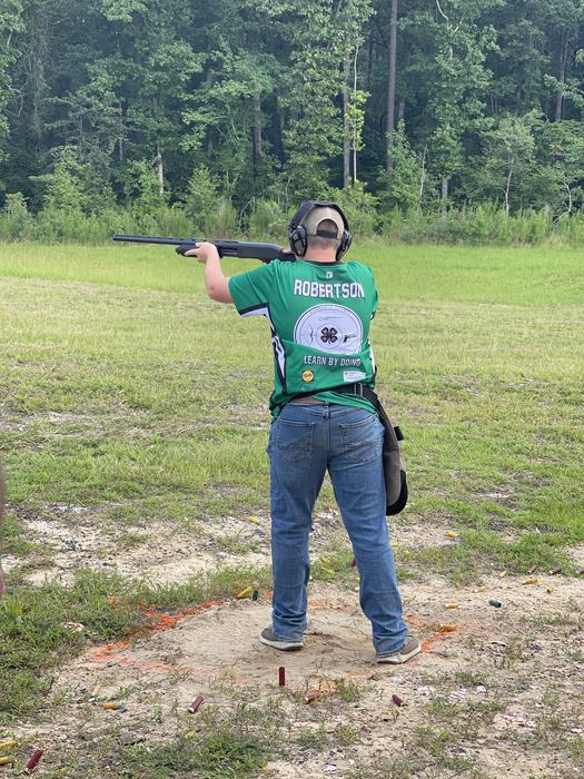 Person wearing green jersey labeled "ROBERTSON" and "LEARN BY DOING" aiming a shotgun at an outdoor range