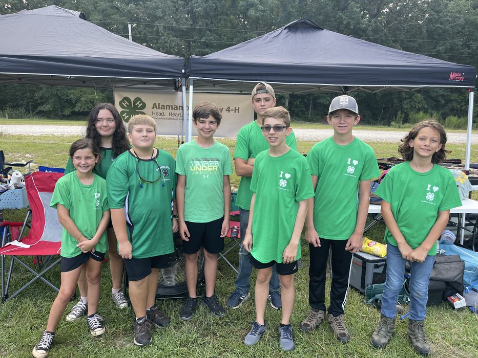 Group of children in green 4-H shirts standing under a canopy with "Alamance County 4‑H" banner.