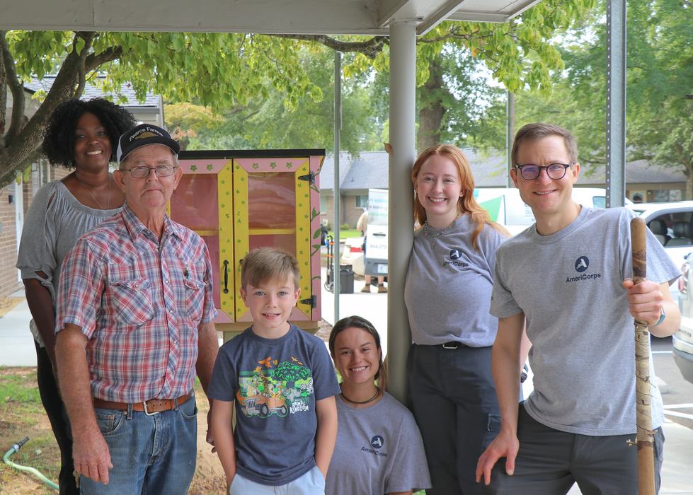 Group of six, including AmeriCorps volunteers and a child, by a small painted pantry