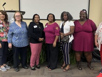 Nine women standing side-by-side in front of a podium and projection screen.