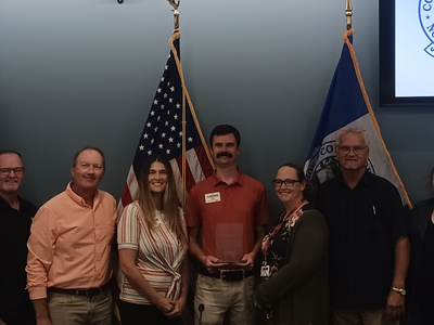 Nine people standing in front of flags, center man holding a clear award plaque.