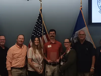 Nine people standing in front of flags, center man holding a clear award plaque.