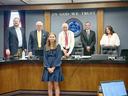 Young girl standing before county commissioners' dais with seal and words "IN GOD WE TRUST"