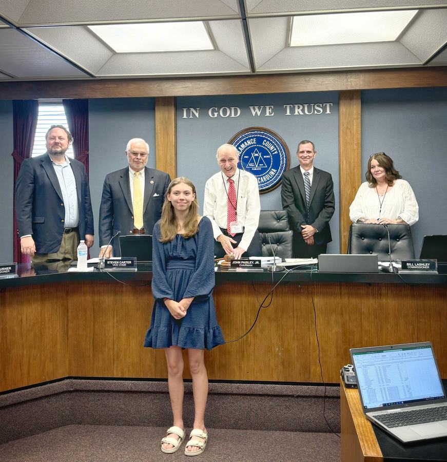 Young girl standing before county commissioners' dais with seal and words "IN GOD WE TRUST"